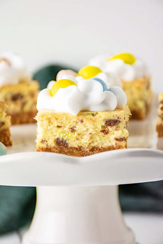 Close-up of Mini Egg Cheesecake Bars with a crumbly base, creamy filling speckled with chocolate chips, and a whipped topping adorned with vibrant yellow and blue candy pieces, all elegantly displayed on a white cake stand.