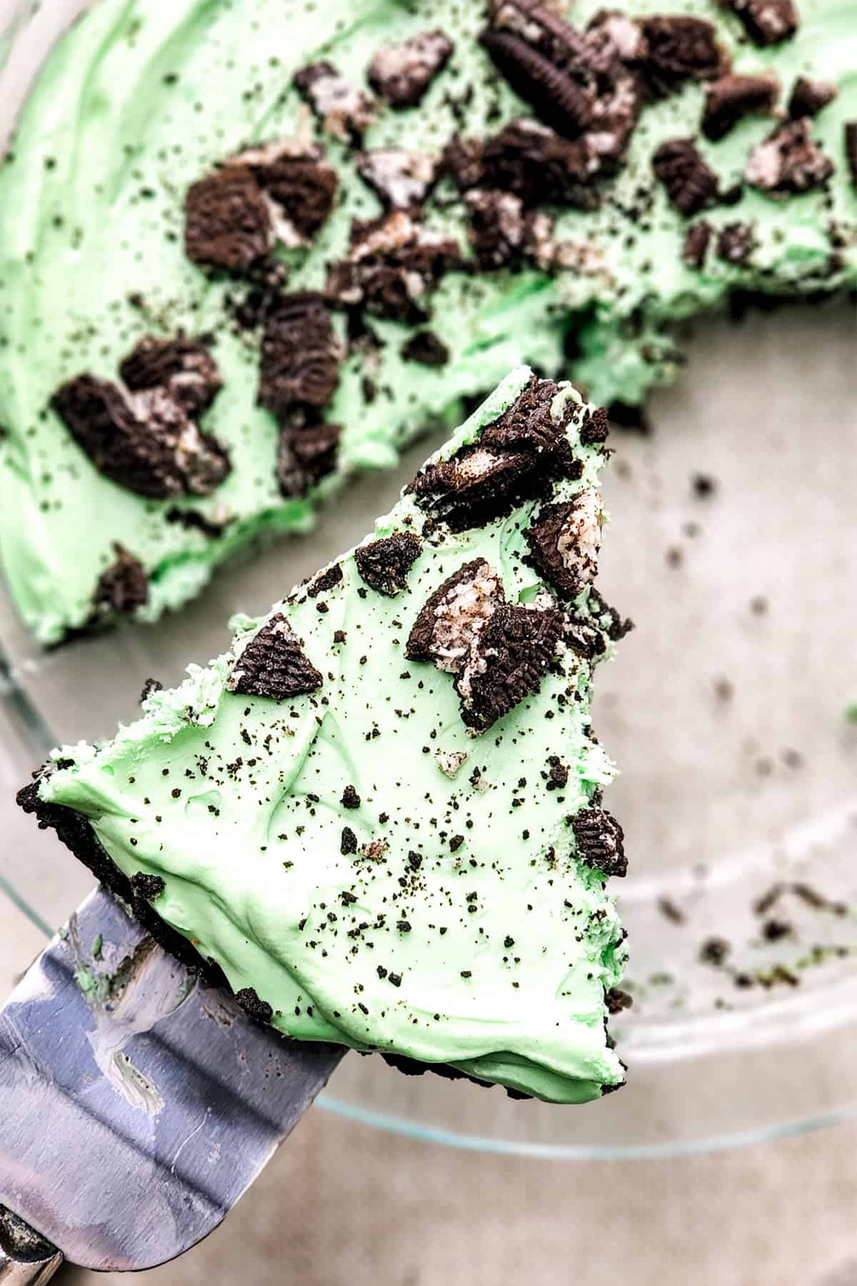 A slice of mint chocolate pie is being served. The pie has a green filling topped with crushed chocolate cookies in a clear dish.