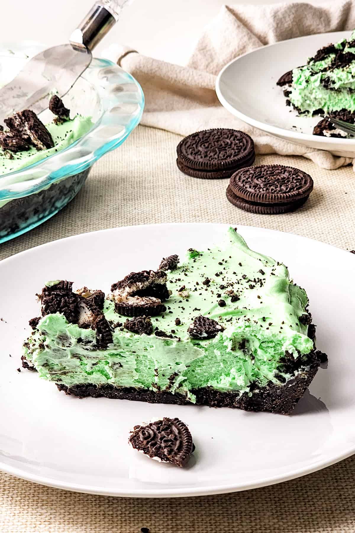 A slice of mint chocolate pie on a plate with crumbled cookies, next to Oreos. A pie server rests on the full pie in the background.