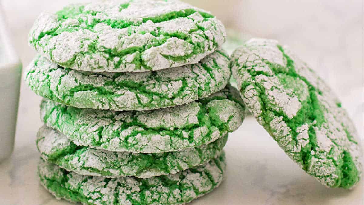 A stack of vibrant green crinkle cookies, crafted from cake mix recipes and dusted with powdered sugar, is elegantly displayed on a white surface.