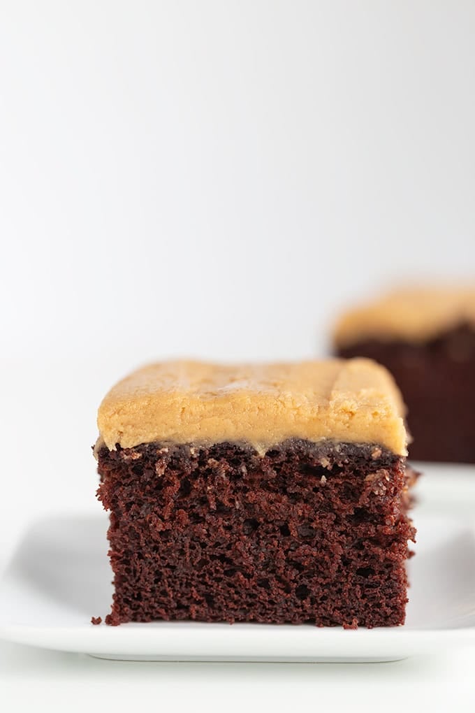 A slice of chocolate cake with creamy peanut butter frosting sits on a white plate, with another slice in the background. This delightful treat is actually a Chocolate Wacky Cake, made without eggs or butter for an unusual twist on a classic favorite.