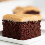A slice of Chocolate Wacky Cake, with a layer of light brown frosting, sits elegantly on a white plate—made without eggs, butter, or milk.