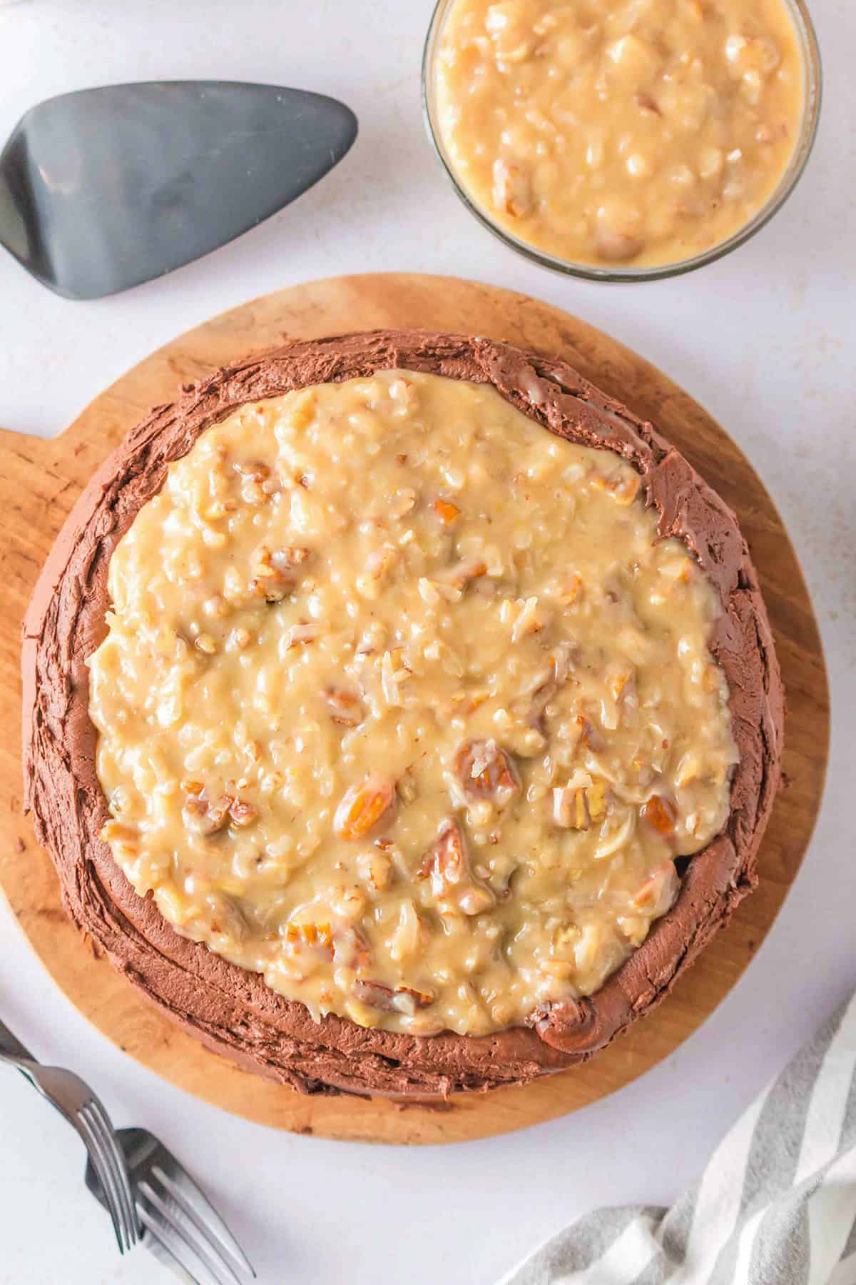 A classic German Chocolate Cake with coconut-pecan frosting sits on a wooden board, accompanied by a bowl of extra frosting, a cake server, and two forks.