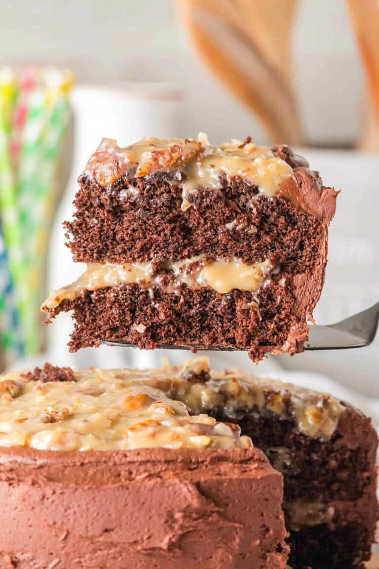 A slice of layered German Chocolate Cake with creamy filling and frosting is lifted from the whole cake, showcasing its rich texture and decadent layers.