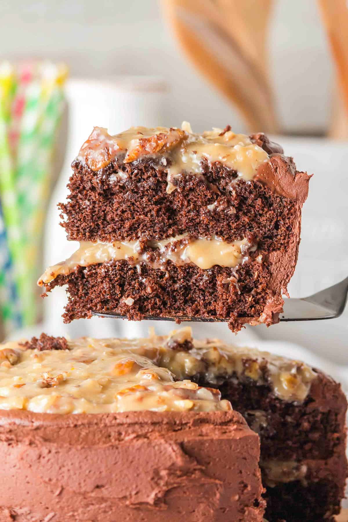 A slice of layered German Chocolate Cake with creamy filling and frosting is lifted from the whole cake, showcasing its rich texture and decadent layers.