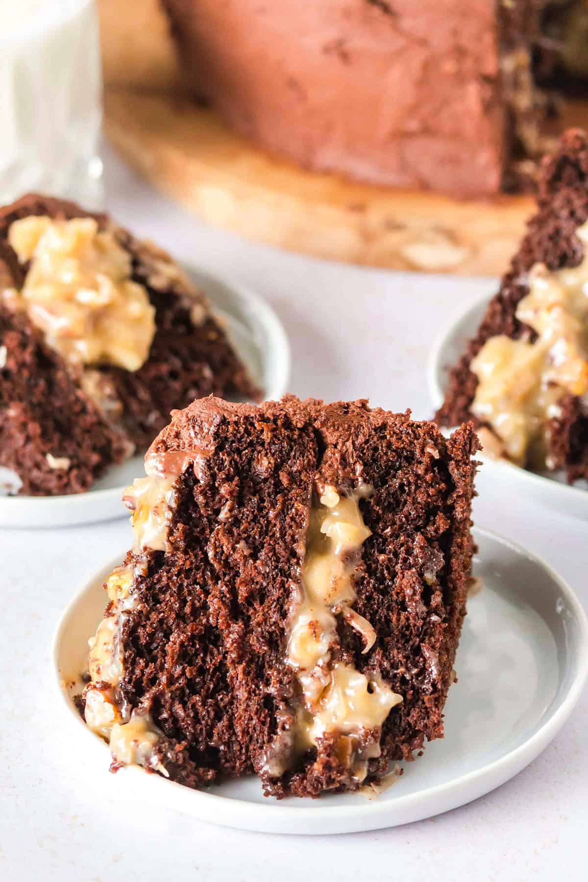 A slice of German Chocolate Cake with a coconut and pecan filling sits on a small white plate, with more slices and the rest of the cake in the background.