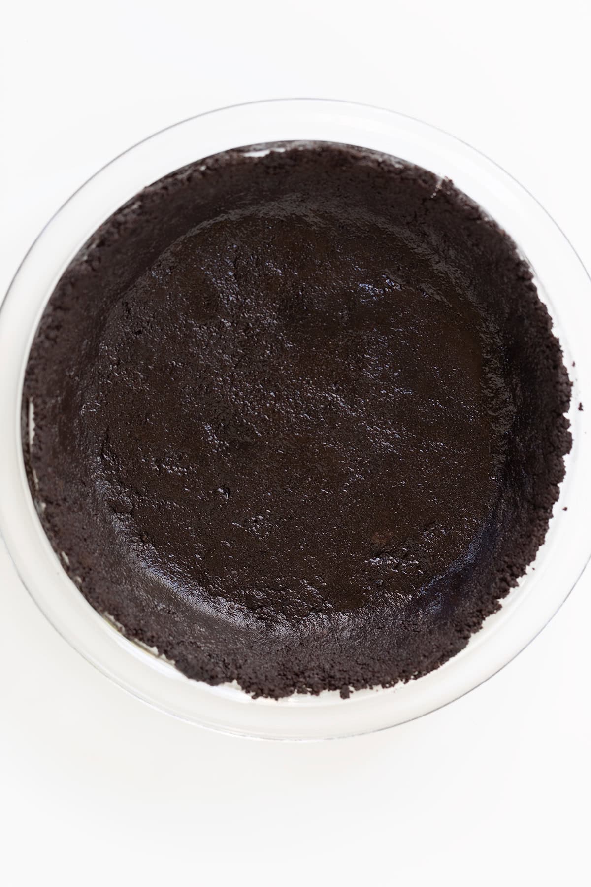 A glass pie dish filled with a pressed chocolate cookie crumb crust, viewed from above on a white surface.
