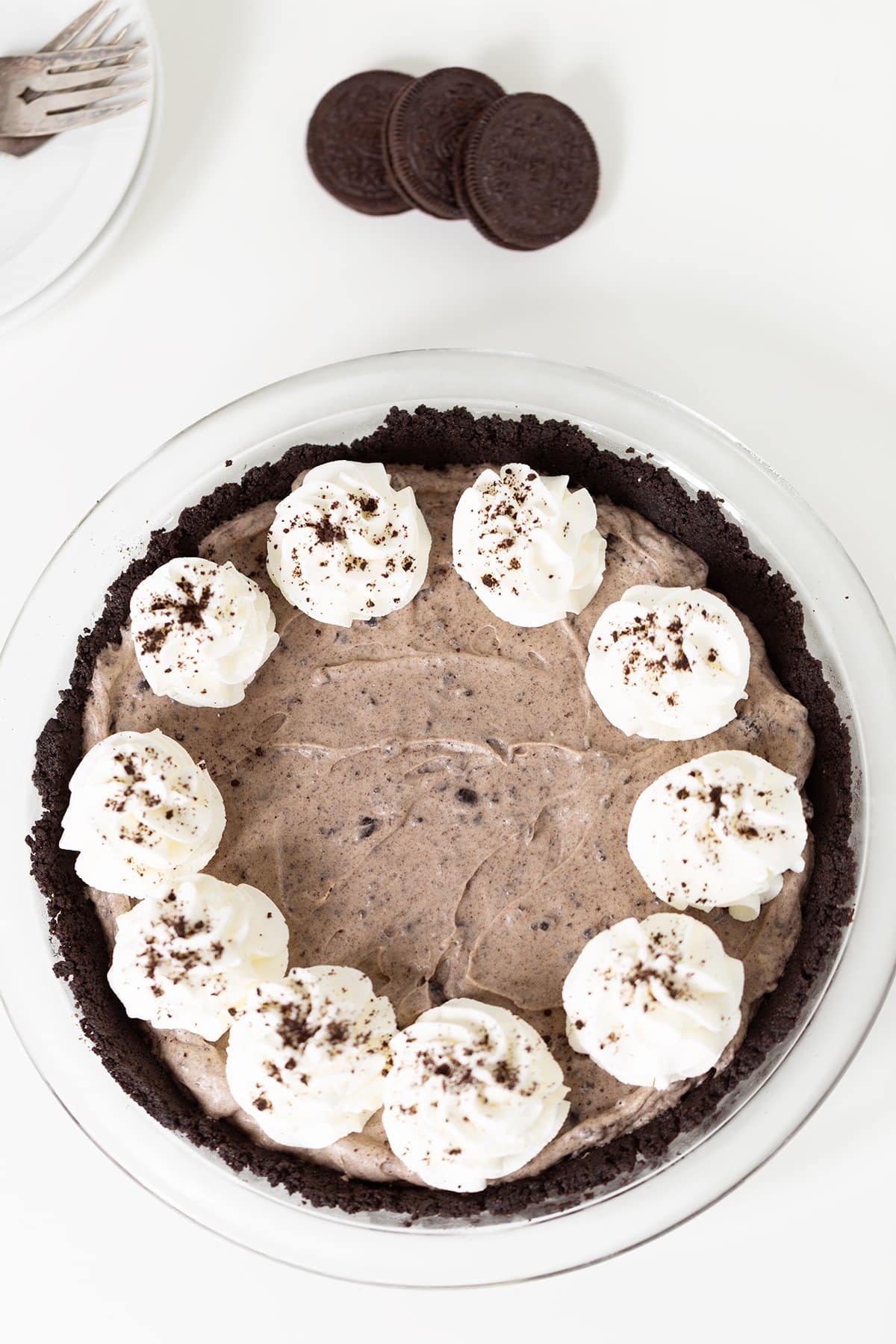 A chocolate cookie crust pie filled with creamy chocolate filling, topped with whipped cream dollops and cookie crumbs, with whole cookies and a plate with forks in the background.