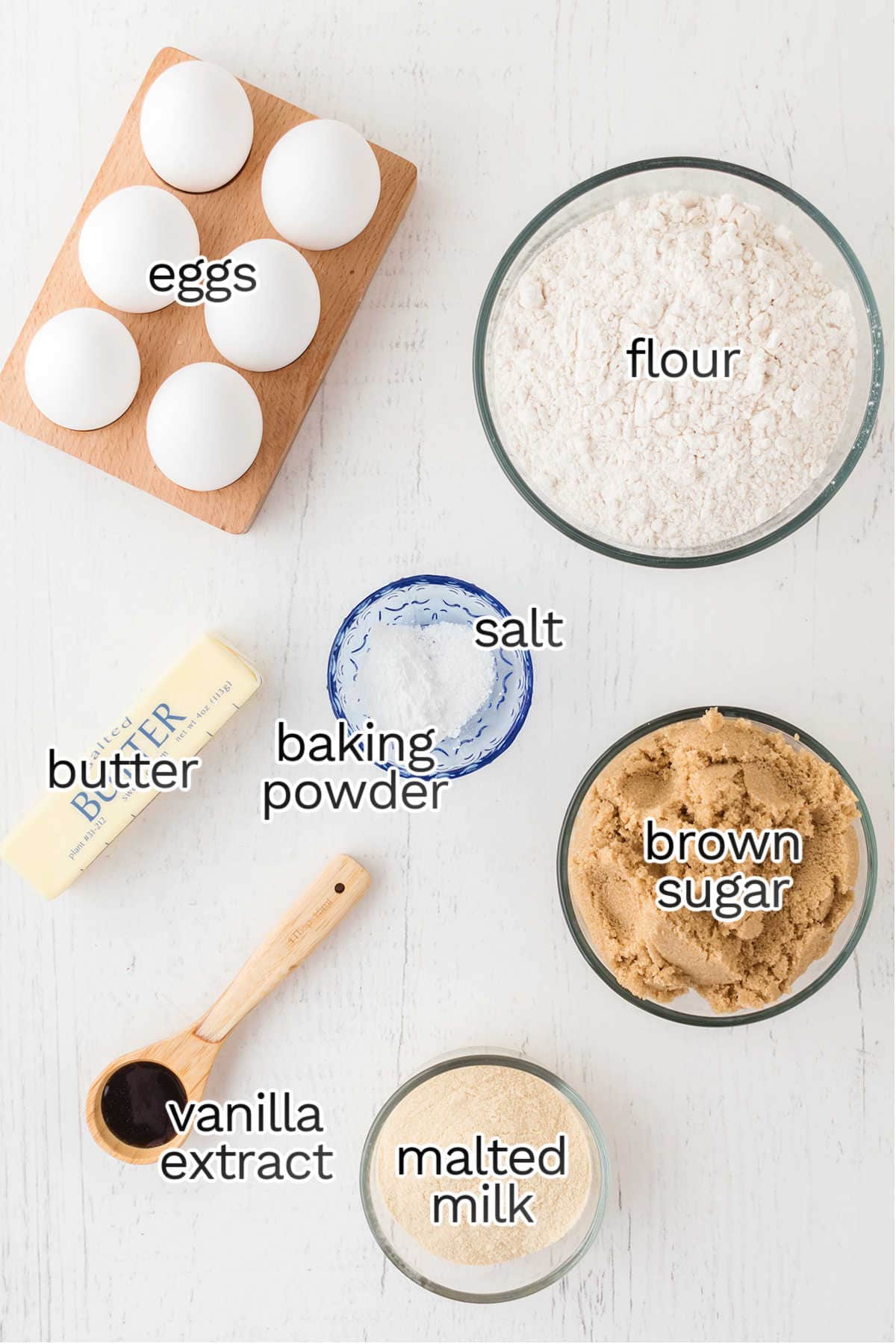 Ingredients for baking blondies on a table: eggs, flour, a butter stick, baking powder, salt, brown sugar, vanilla extract, and malted milk.