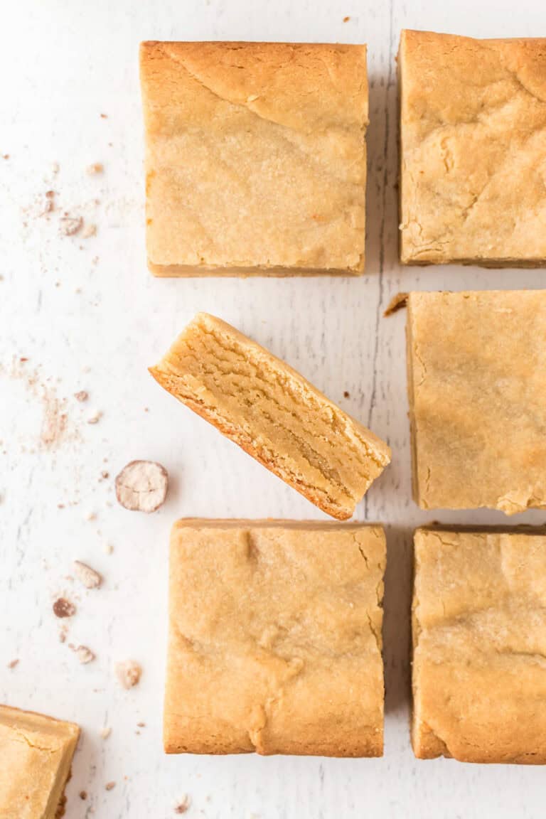 Six square malted milk blondies on a white surface, one turned on its side revealing the interior texture. Crumbs scattered around.