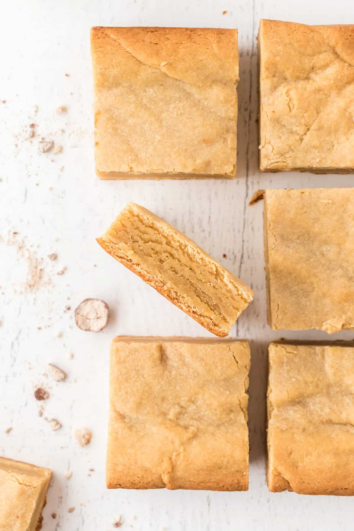 Six square malted milk blondies on a white surface, one turned on its side revealing the interior texture. Crumbs scattered around.