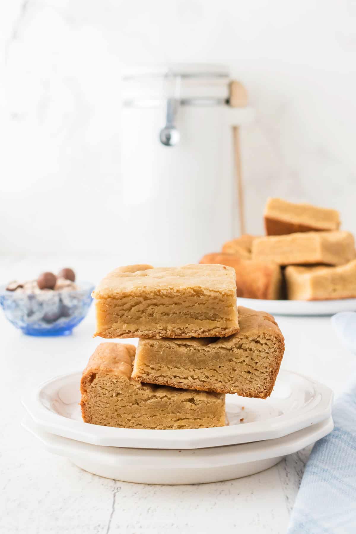 Three stacked blondies sit on a plate, with a jar and a bowl of mixed nuts in the background, exuding a hint of malted milk for an irresistible twist.