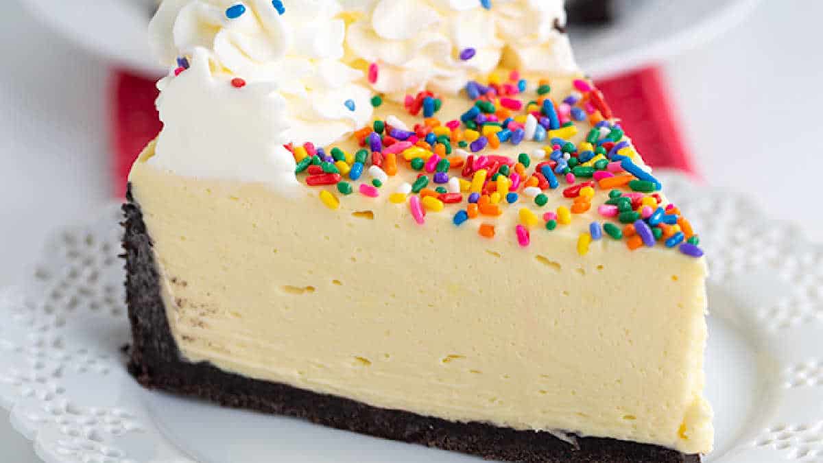 A slice of cheesecake, straight from the realm of delicious cake ideas, is topped with colorful sprinkles and whipped cream, served on a white doily-lined plate.