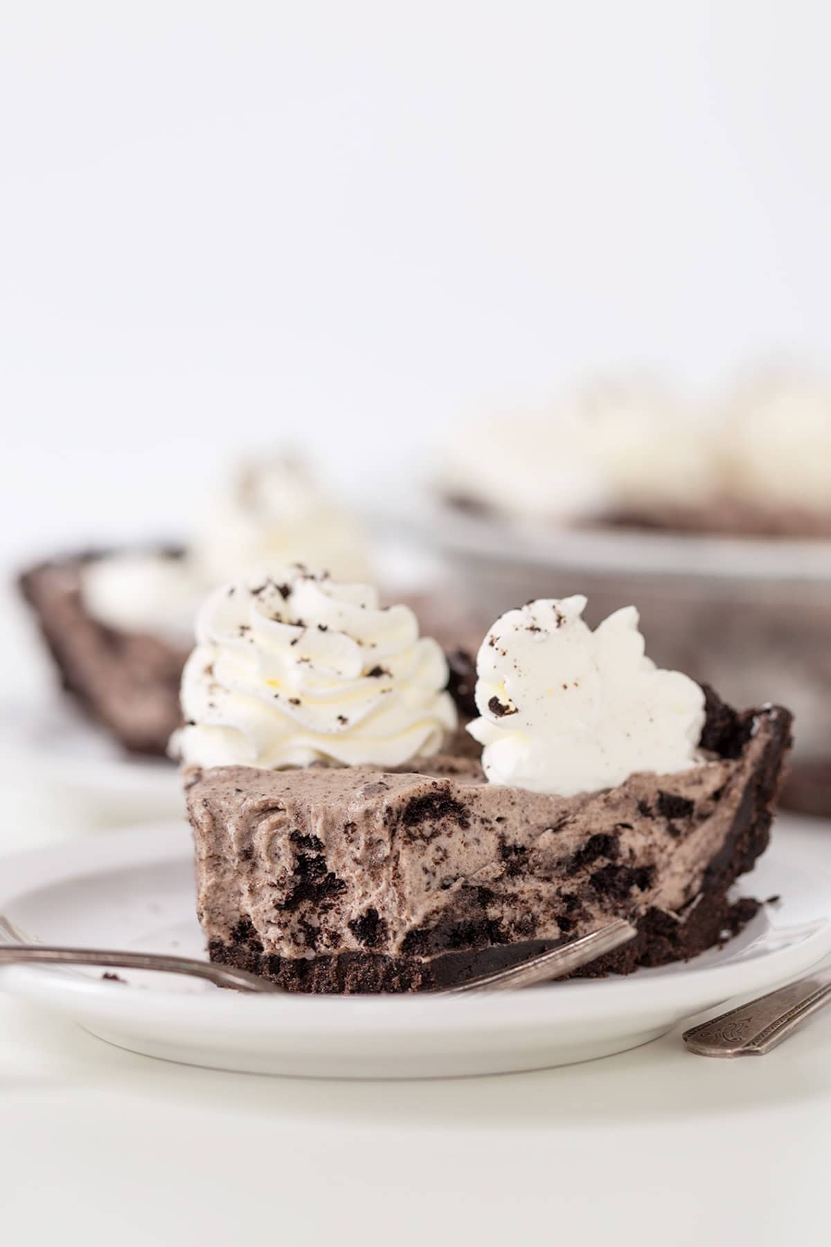 A slice of cookies and cream pie with whipped cream on a plate, a fork beside it, and the remaining pie blurred in the background.