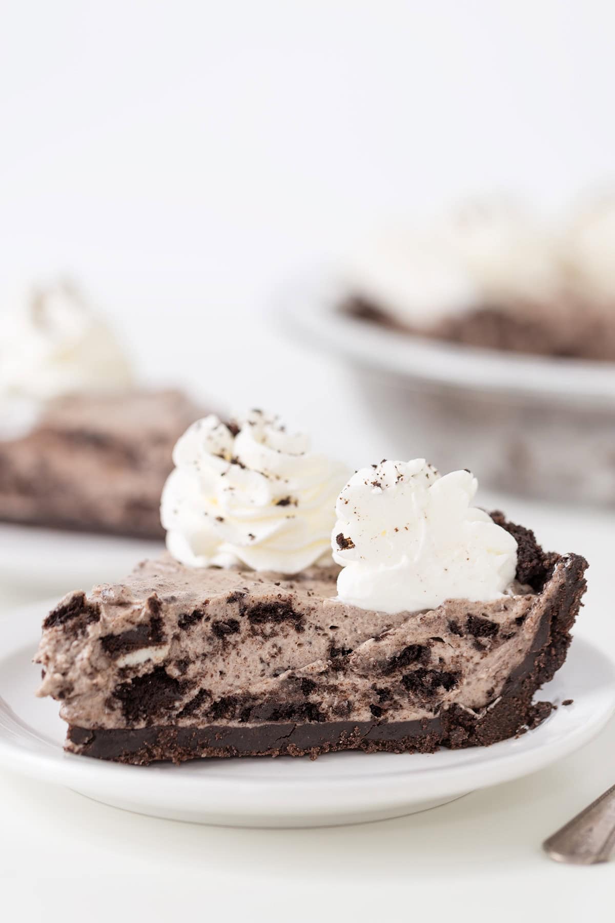 A slice of cookies and cream pie with a chocolate crust, topped with whipped cream, on a white plate.