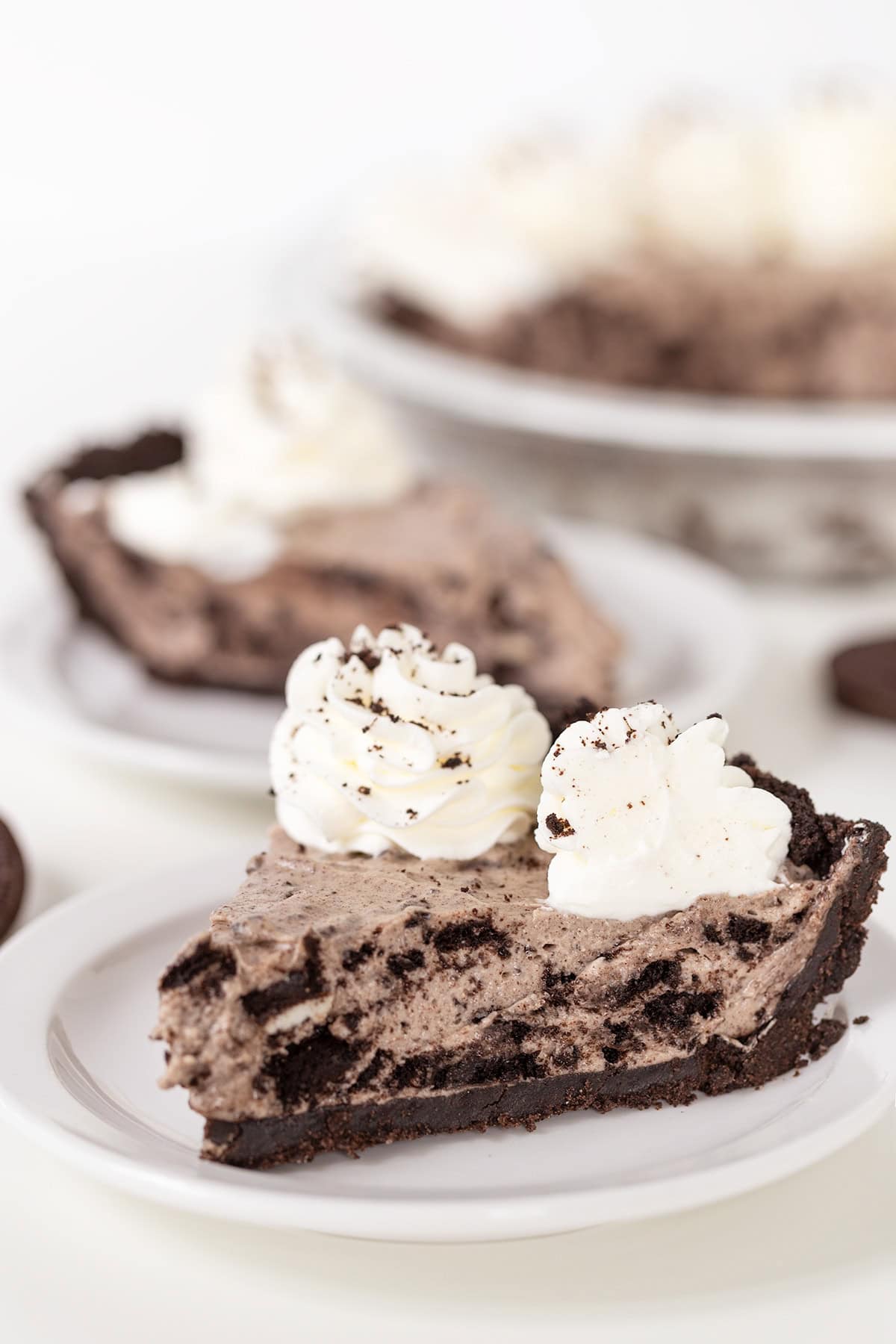 A slice of cookies and cream pie with chocolate crust and whipped cream topping on a white plate, with more slices and a whole pie in the background.