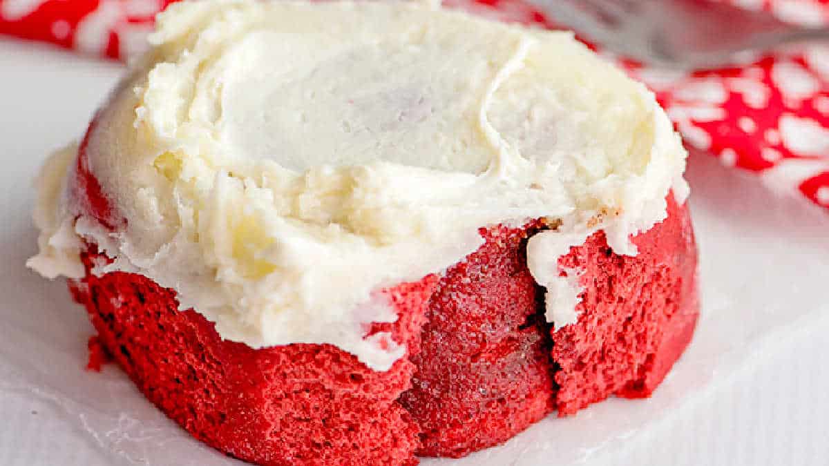 Indulge in a red velvet cinnamon roll, enhanced with cake mix and topped with creamy white frosting, all resting on parchment paper.
