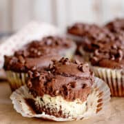 Chocolate cheesecake muffins with a marbled chocolate and vanilla interior, one partially unwrapped and missing a bite, sitting on brown parchment paper.
