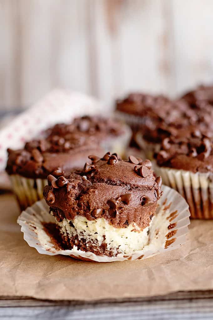 Chocolate cheesecake muffins with a marbled chocolate and vanilla interior, one partially unwrapped and missing a bite, sitting on brown parchment paper.