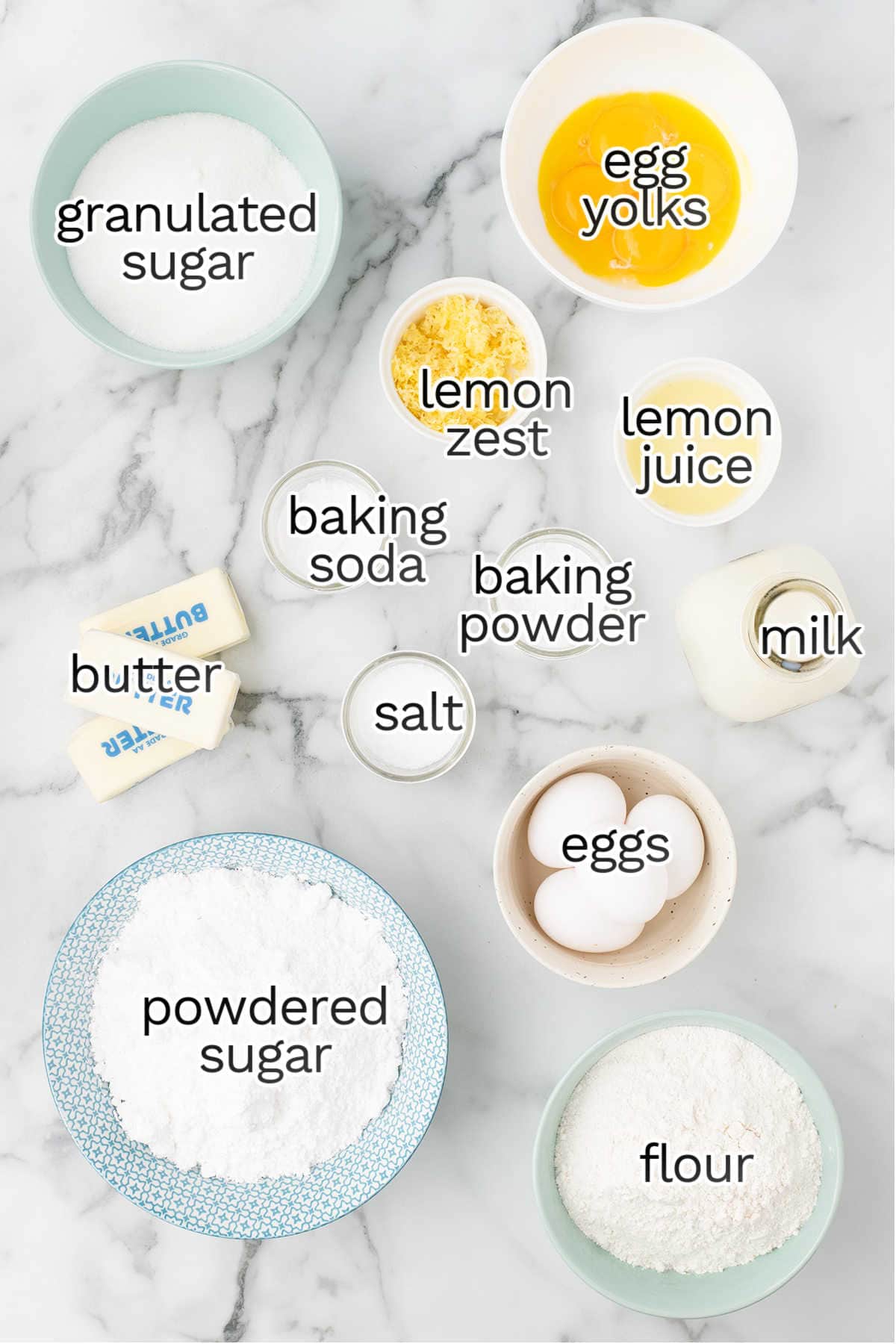 Baking ingredients for Lemon Cake on a marble countertop, including granulated sugar, powdered sugar, flour, butter, eggs, egg yolks, lemon zest, lemon juice, baking soda, baking powder, salt, and milk.