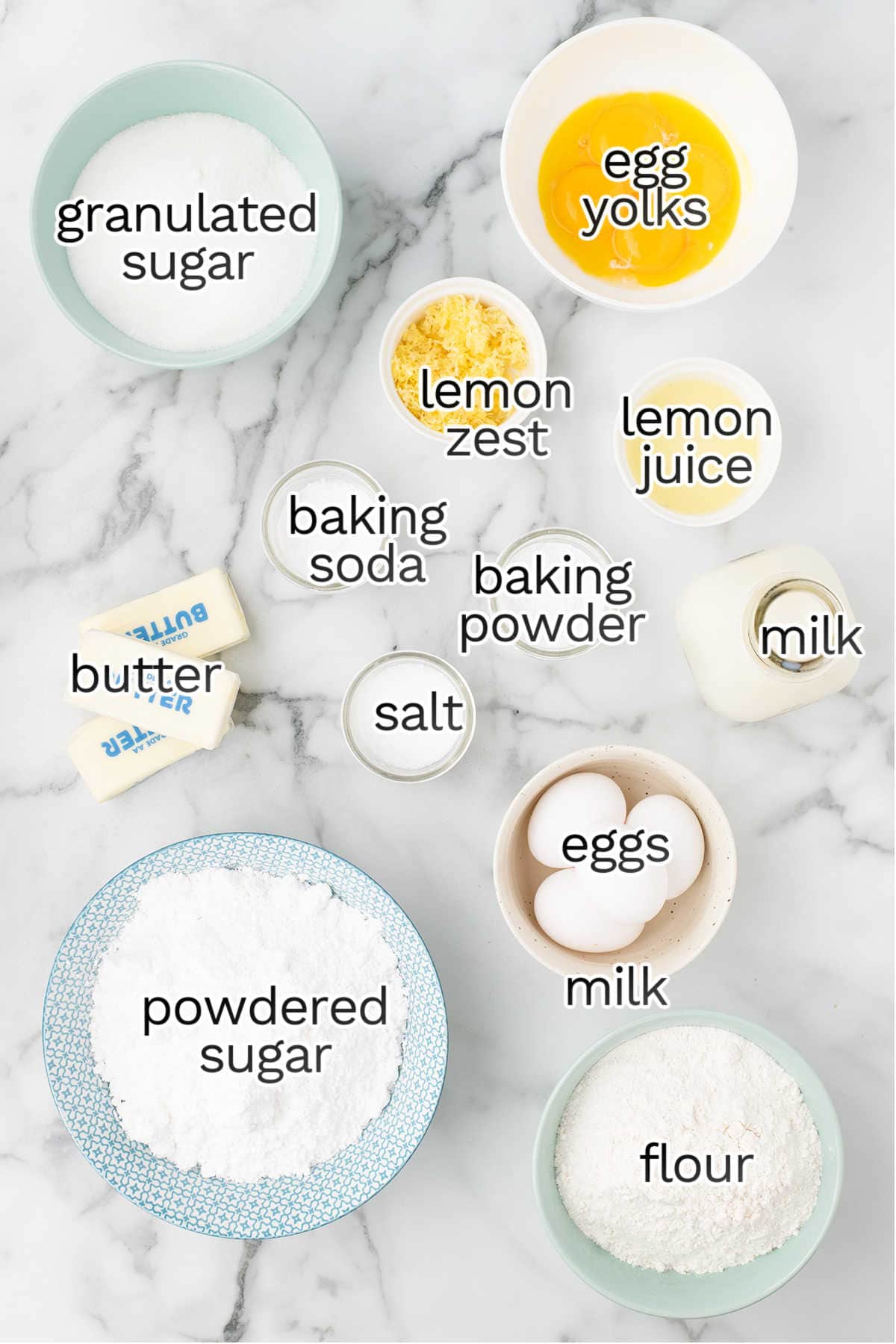 Baking ingredients arranged on a marble countertop, including eggs, egg yolks, flour, milk, butter, granulated sugar, powdered sugar, lemon zest, lemon juice, baking soda, baking powder, and salt.