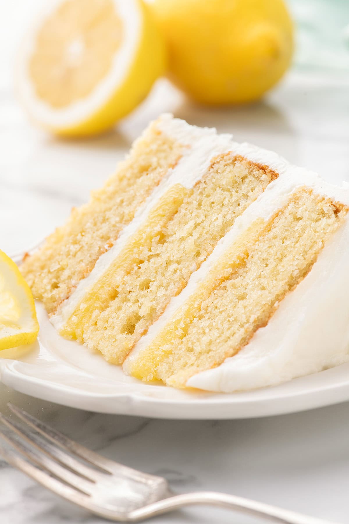 A slice of three-layer lemon cake with white frosting and lemon filling on a white plate, with a fork and lemon halves in the background.