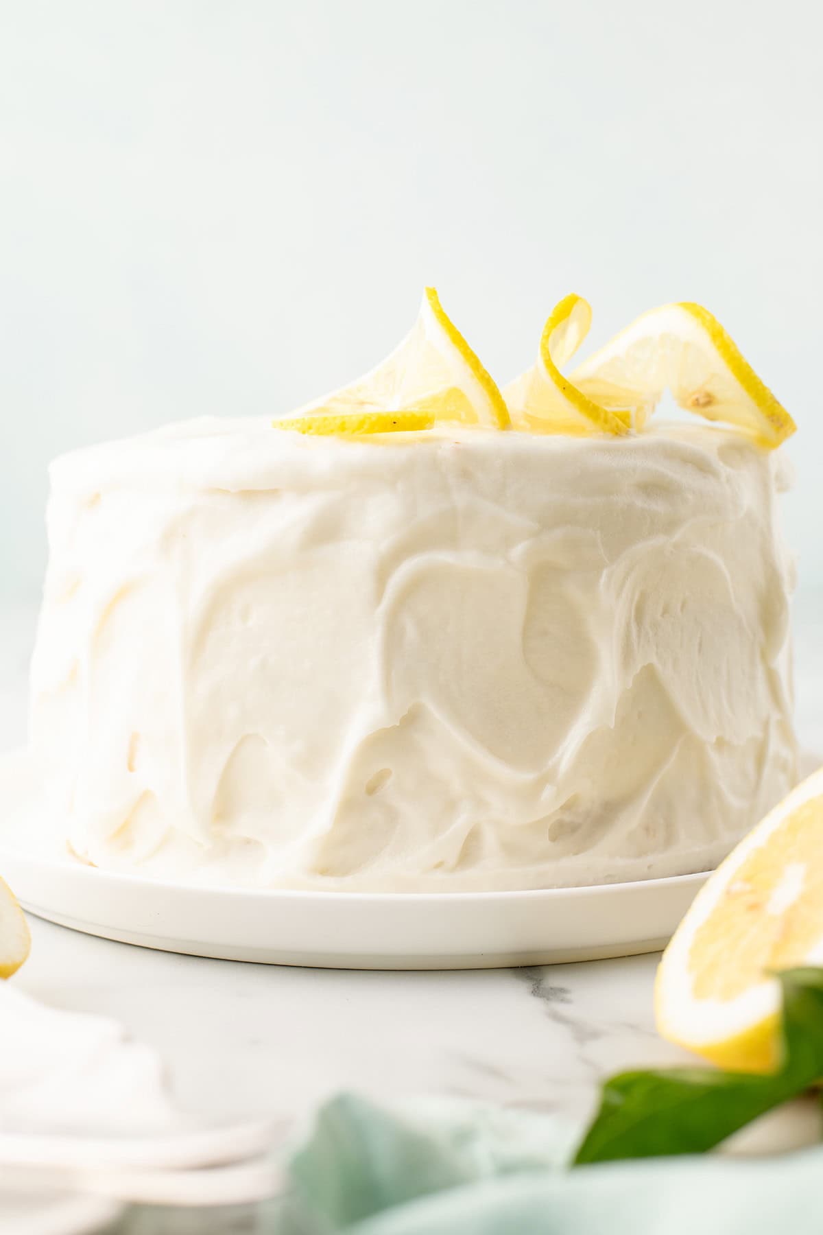 A round frosted cake sits on a white plate, decorated with lemon slices and lemon zest curls on top.