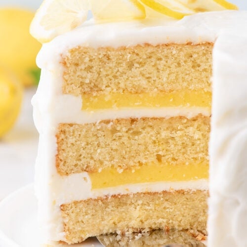 A three-layer lemon cake with white icing and lemon slices on top, shown with a slice being removed.