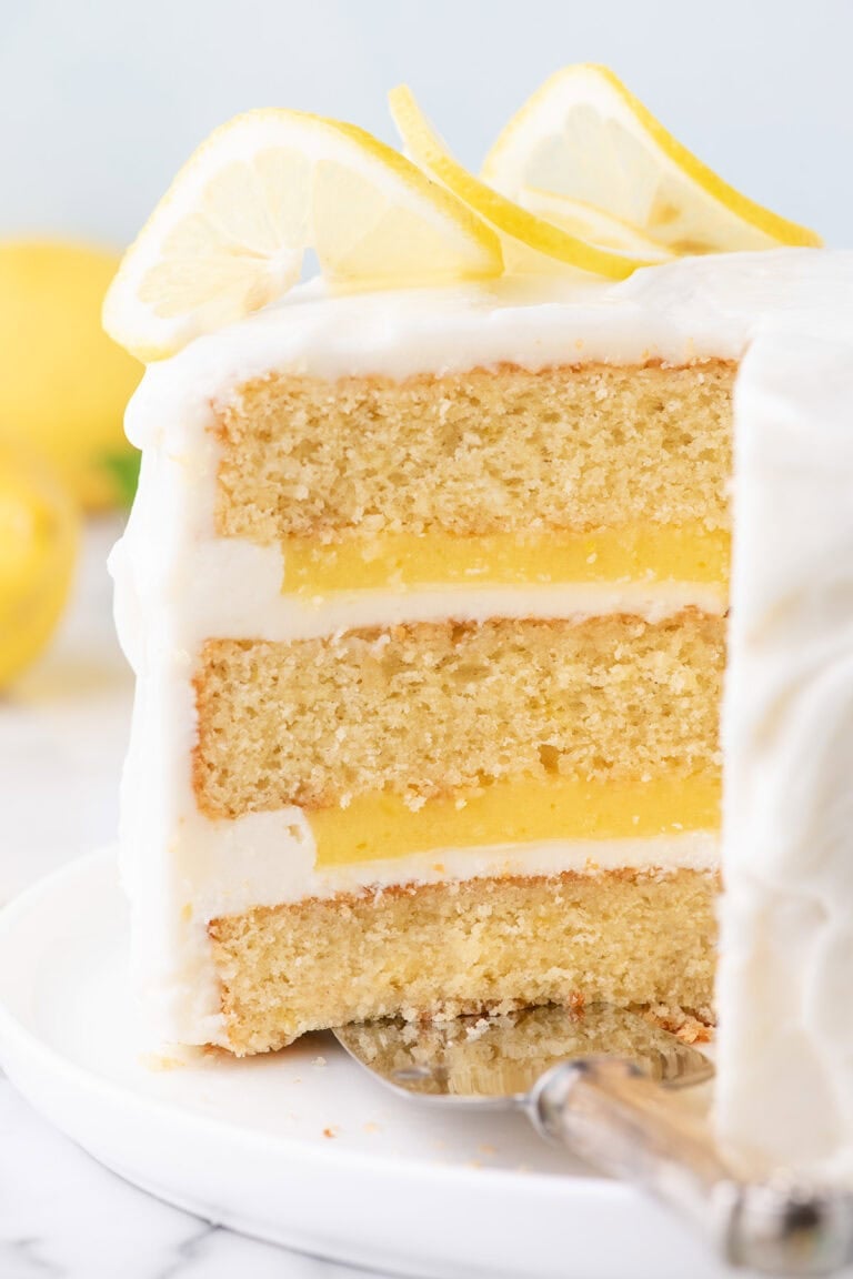 A three-layer lemon cake with white icing and lemon slices on top, shown with a slice being removed.
