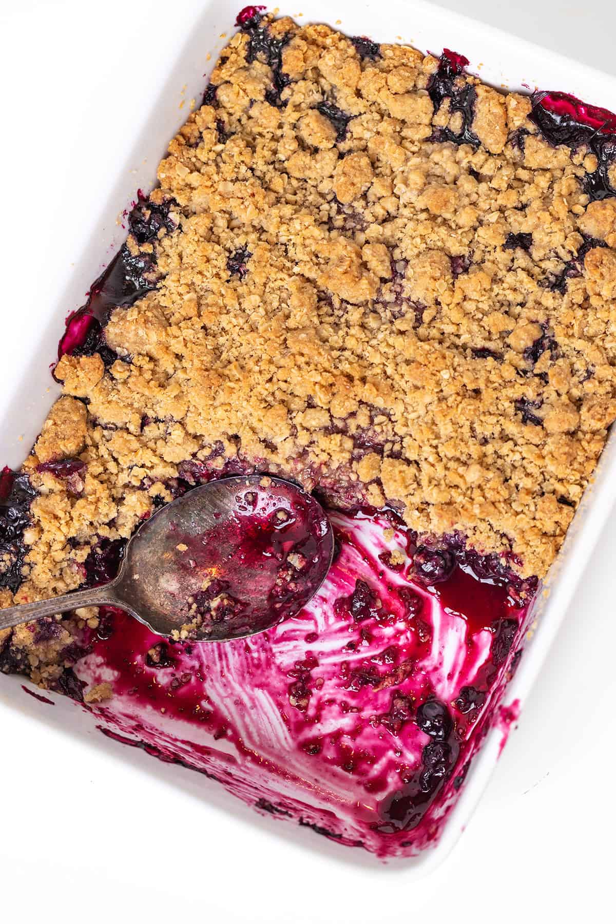 Baking dish with partially eaten Blueberry Crisp, showing a metal spoon and vibrant berry filling beneath a golden crumb topping.