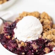 A serving of Blueberry Crisp topped with a scoop of vanilla ice cream on a white plate, with a spoon resting beside it.