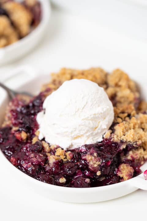 A serving of Blueberry Crisp topped with a scoop of vanilla ice cream on a white plate, with a spoon resting beside it.
