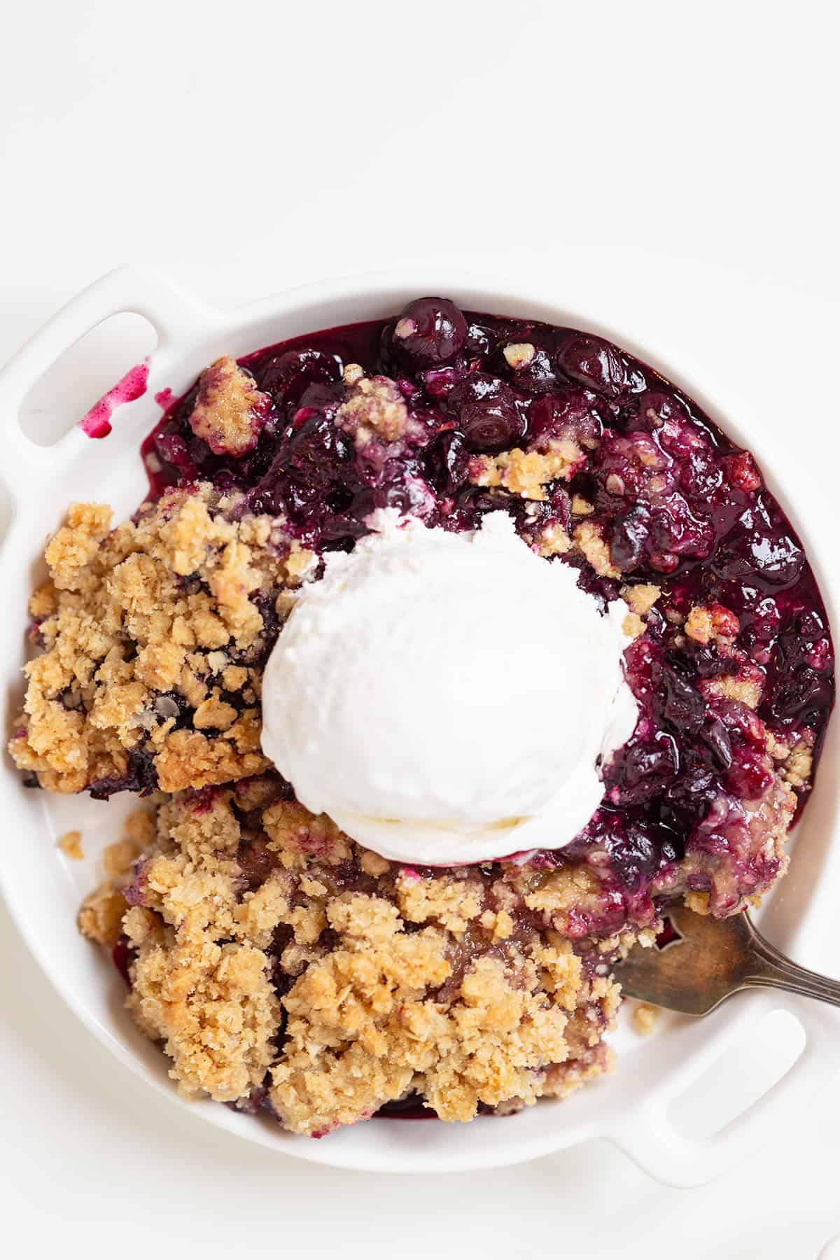 A white dish with Blueberry Crisp crumble topped with a scoop of vanilla ice cream and a spoon on the side.
