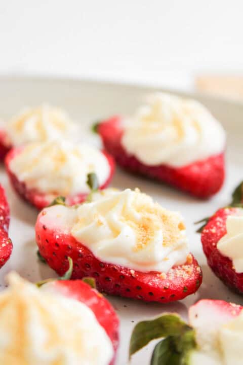 Halved strawberries topped with a swirl of cream and sprinkled with cookie crumbs are arranged on a white plate, creating irresistible Cheesecake Strawberries.