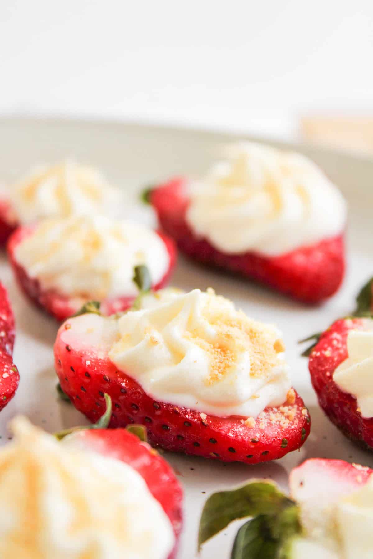 Halved strawberries topped with a swirl of cream and sprinkled with cookie crumbs are arranged on a white plate, creating irresistible Cheesecake Strawberries.