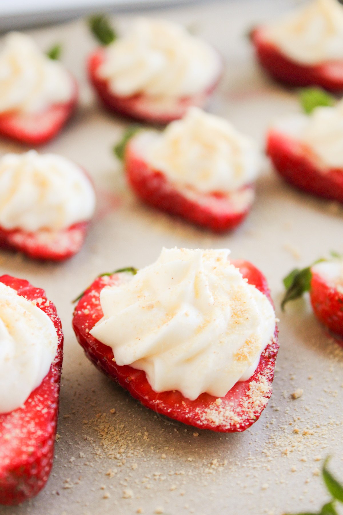 Halved strawberries topped with piped cream and a light dusting of crumbs, these Deviled Strawberries are arranged on a light-colored surface for a delightful cheesecake-inspired treat.
