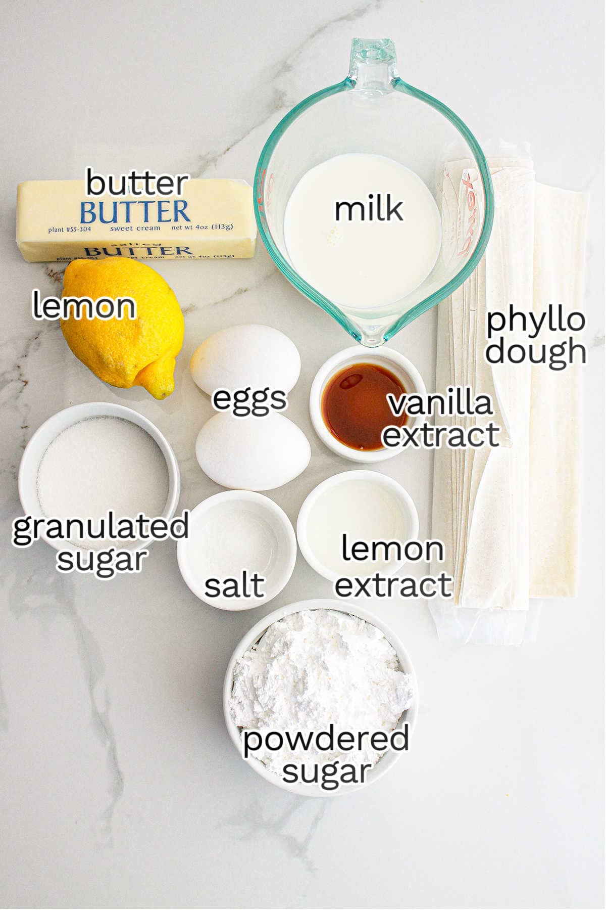 Various ingredients for a recipe are arranged on a white surface, including butter, milk, phyllo dough—perfect for making lemon crinkle cake—lemon, eggs, vanilla and lemon extracts, sugar, salt, and powdered sugar.