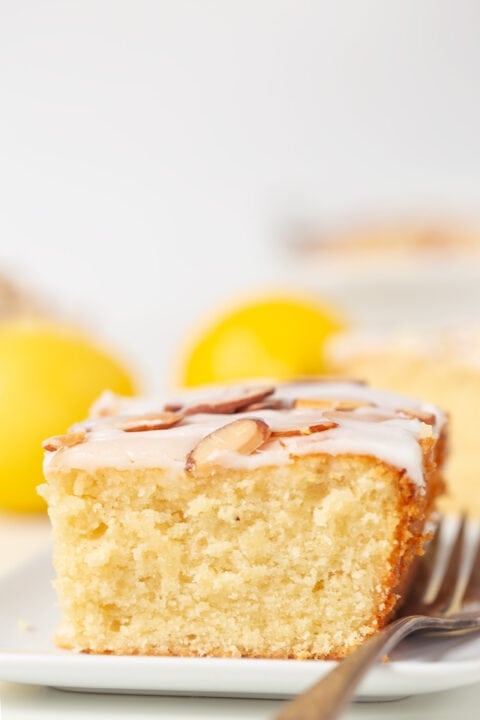 A slice of Lemon Almond Cake with almond slices on top, placed on a white plate with a fork; two yellow lemons are in the background.