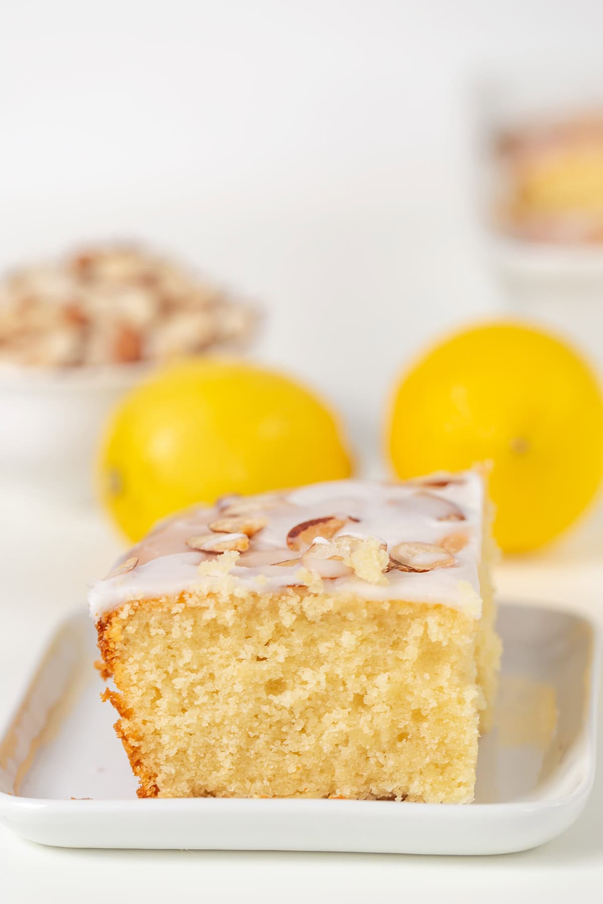A slice of Lemon Almond Cake with white icing and almond slices sits on a small plate, with two whole lemons and a bowl of chopped nuts in the background.