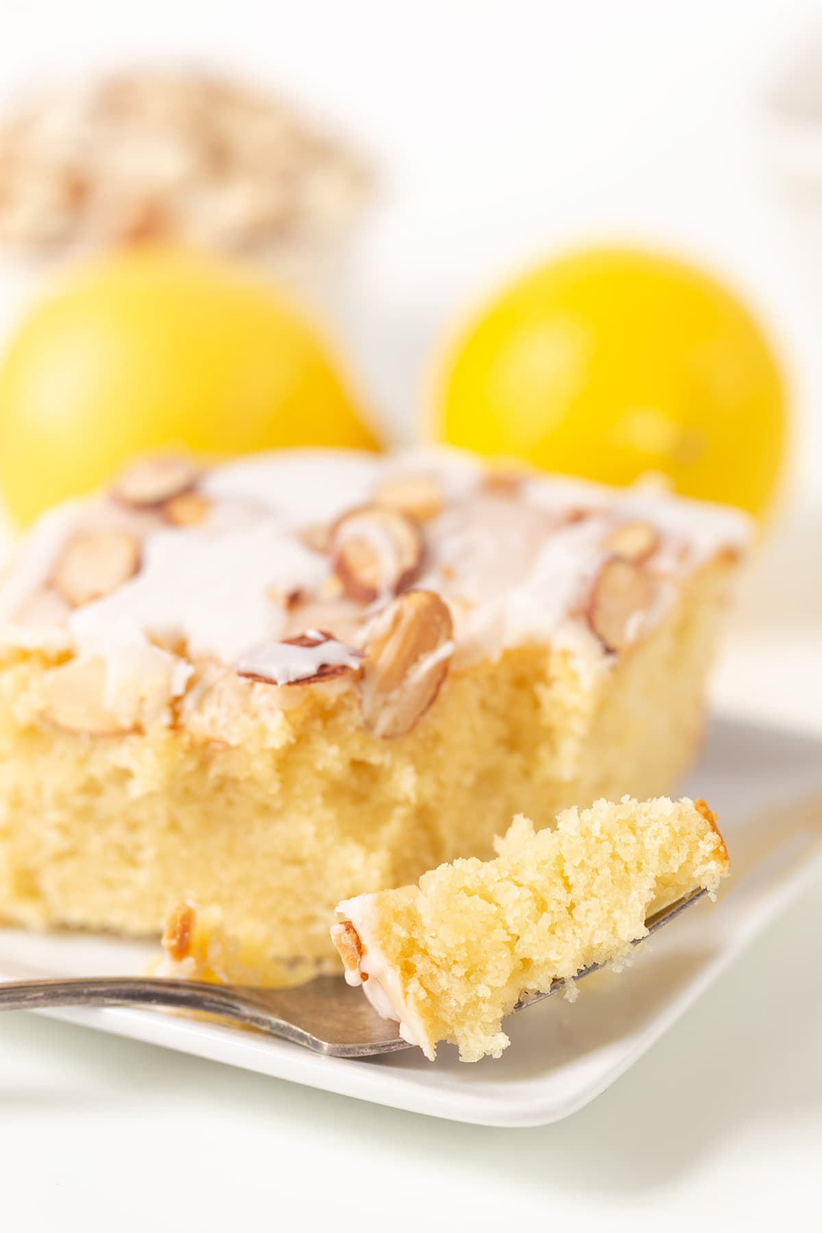 A slice of Lemon Almond Cake with a piece on a fork sits on a white plate, accompanied by two whole lemons in the background.
