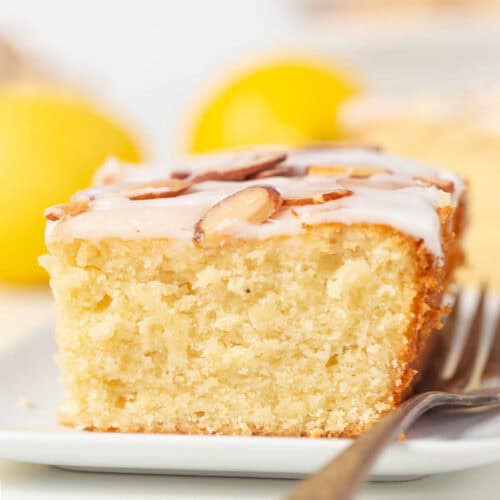 A square slice of Lemon Almond Cake with white icing and sliced almonds on top, served on a white plate with a fork. Lemons are blurred in the background.