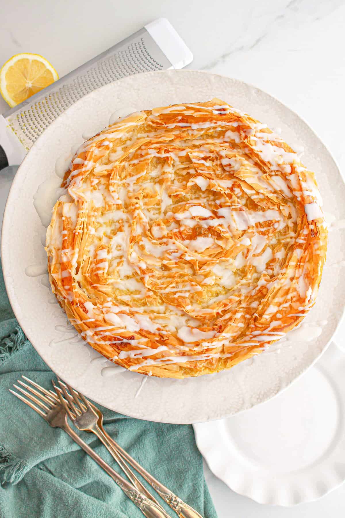 A round, golden-brown Lemon Crinkle Cake drizzled with white icing is displayed on a white plate, next to silver forks, a napkin, and a sliced lemon.