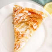 A slice of flaky phyllo dough pastry with icing drizzle sits on a white plate, with a lemon slice and green cloth in the background.