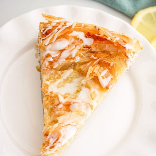 A slice of flaky phyllo dough pastry with icing drizzle sits on a white plate, with a lemon slice and green cloth in the background.