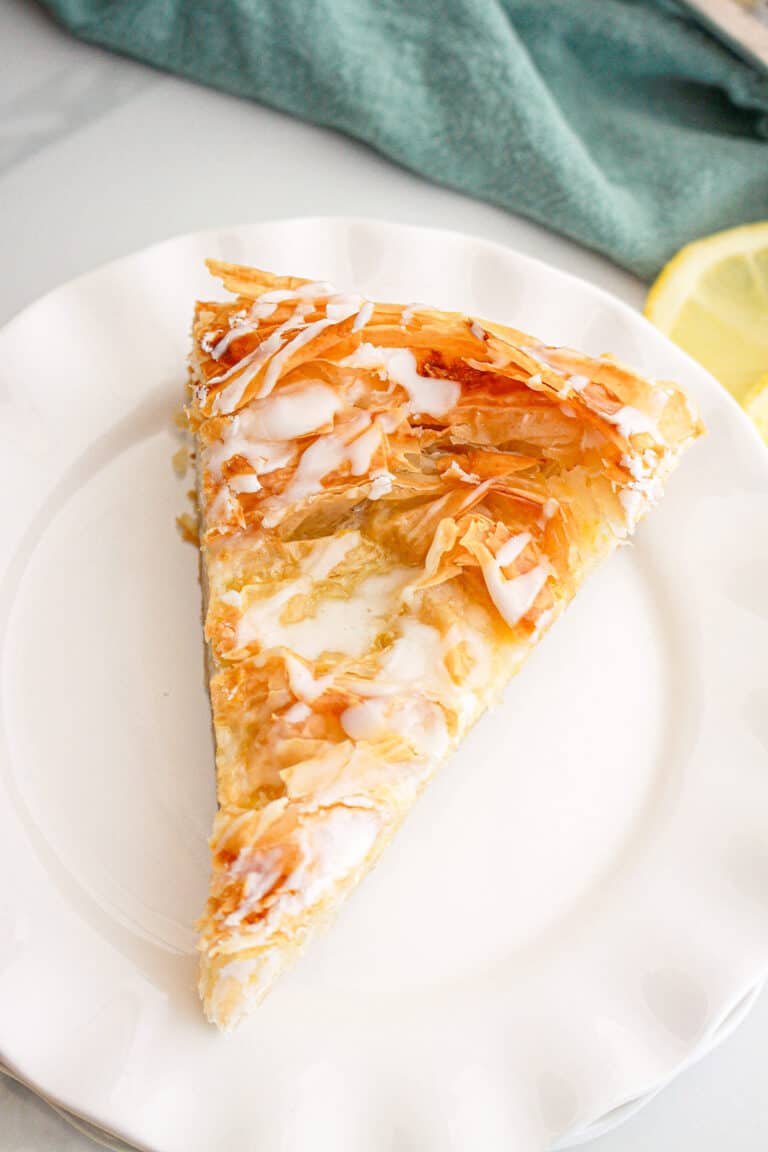 A slice of flaky phyllo dough pastry with icing drizzle sits on a white plate, with a lemon slice and green cloth in the background.