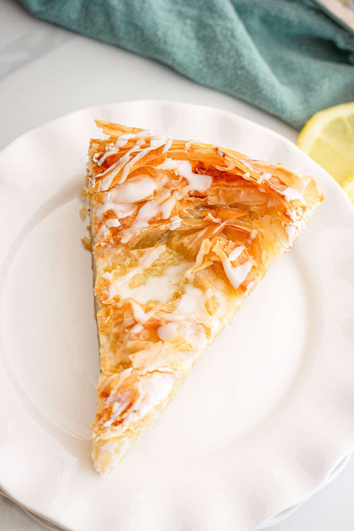 A slice of flaky phyllo dough pastry with icing drizzle sits on a white plate, with a lemon slice and green cloth in the background.