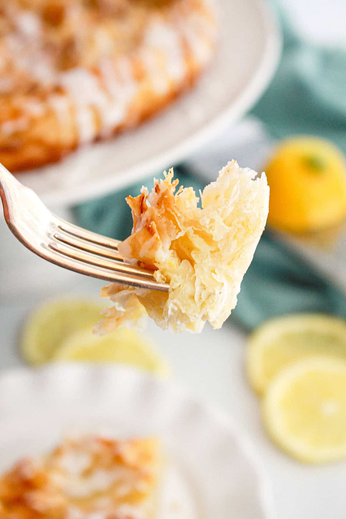 A close-up of a fork holding a bite of flaky phyllo dough pastry with icing, with lemon slices and a blurred Lemon Crinkle Cake in the background.