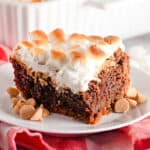 A slice of chocolate cake topped with toasted marshmallows sits on a white plate, surrounded by butterscotch chips—reminiscent of decadent Peanut Butter S'mores Brownies.