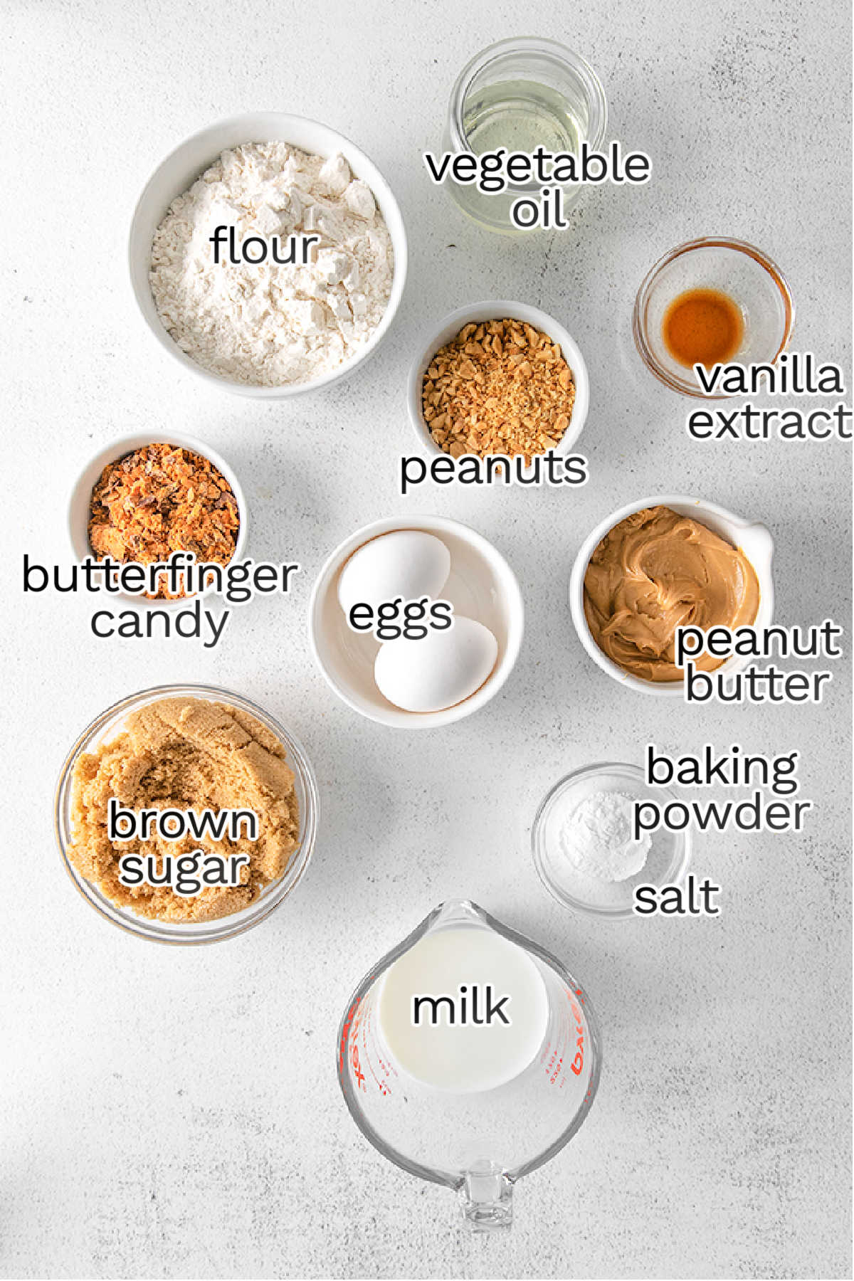 A flat lay of ingredients for baking peanut butter cupcakes, including flour, vegetable oil, vanilla extract, peanuts, peanut butter, eggs, Butterfinger candy, brown sugar, baking powder, salt, and milk.