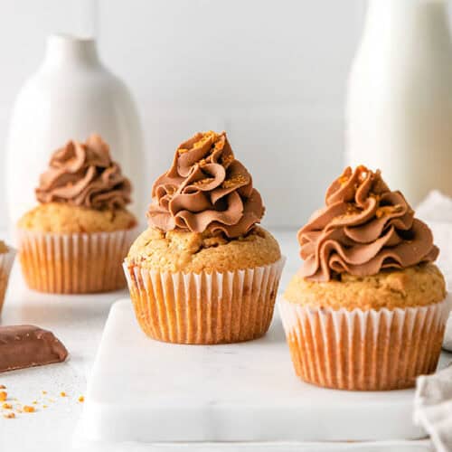 Three peanut butter cupcakes with chocolate frosting are arranged on a marble surface, with a white vase and milk bottle in the background.
